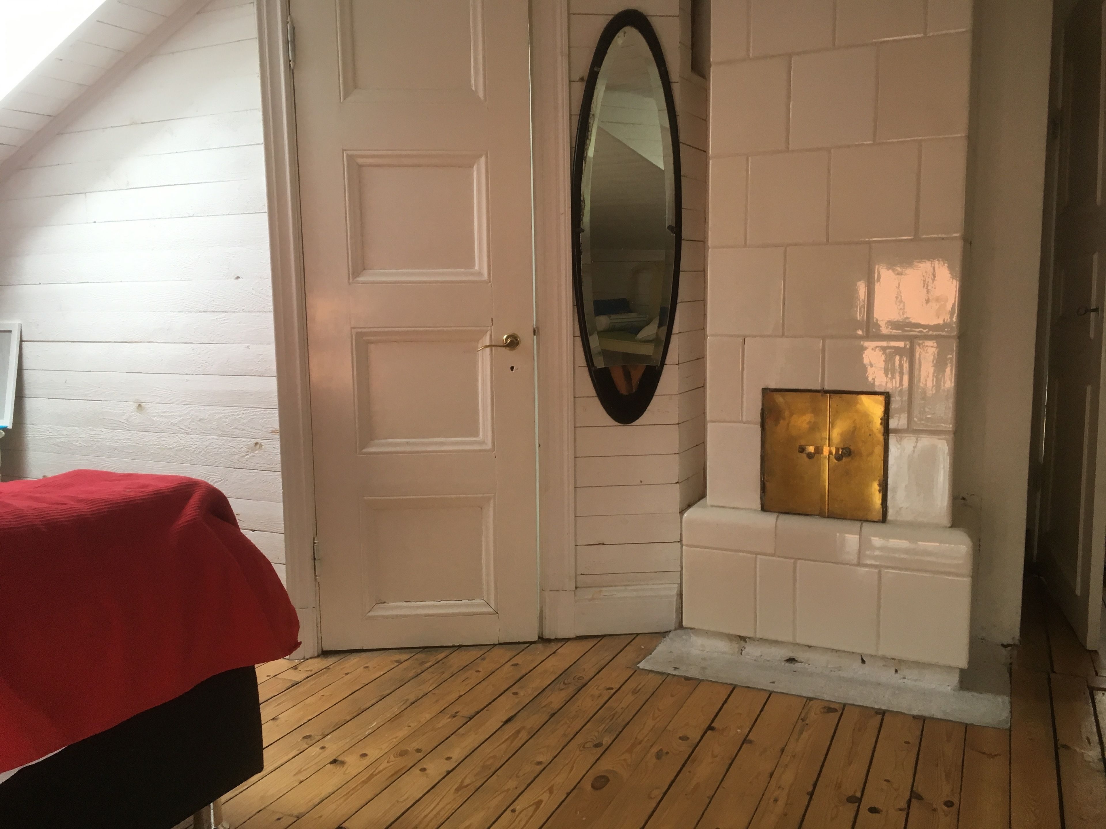 A room with a white door, a mirror, a small fireplace, and a red bedspread.