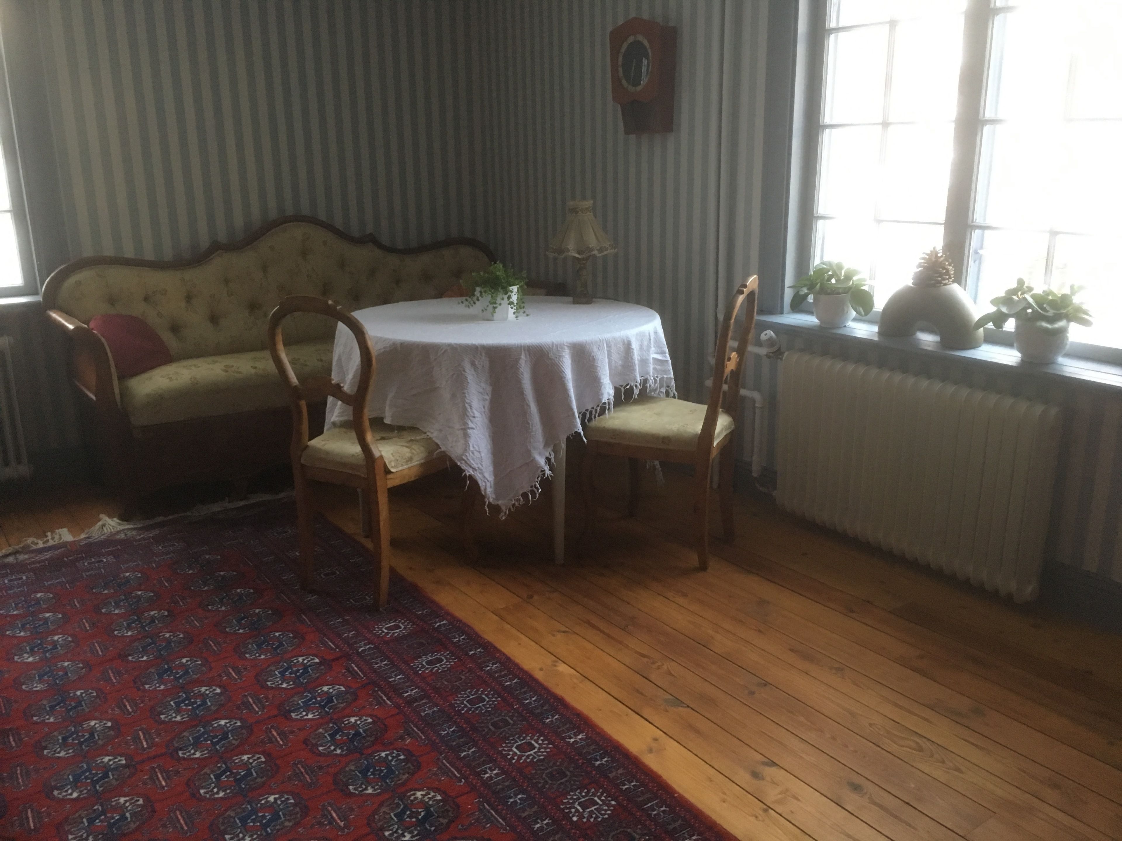 Et værelse med et rundt bord dækket af et hvidt linned, to træstole, en tæppe med et mønster, en sofa, en radiator og et vindue med planter.