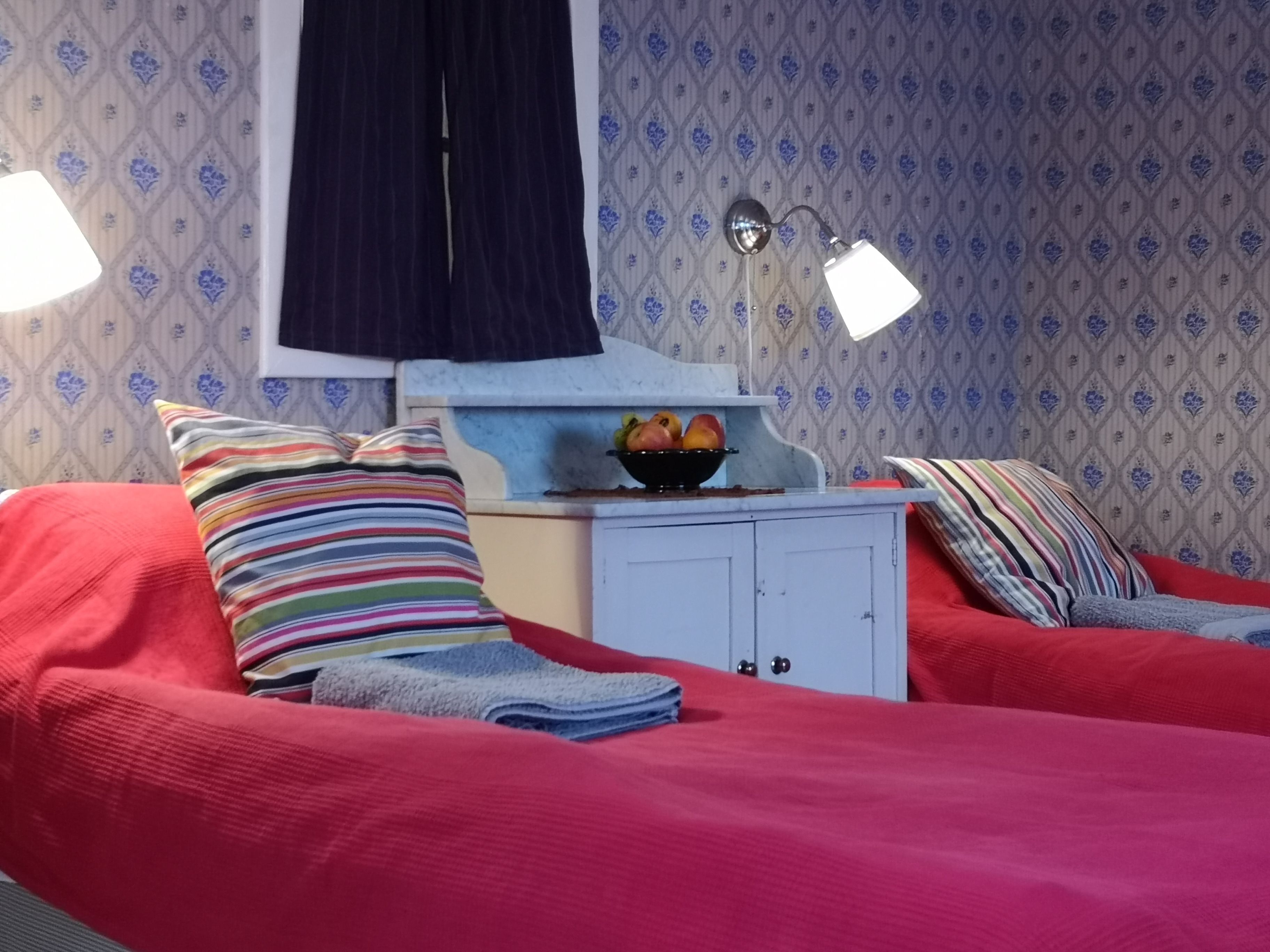 Un dormitorio con dos camas, un armario blanco pequeño, una ventana con cortina oscura y una pared con estampado floral.