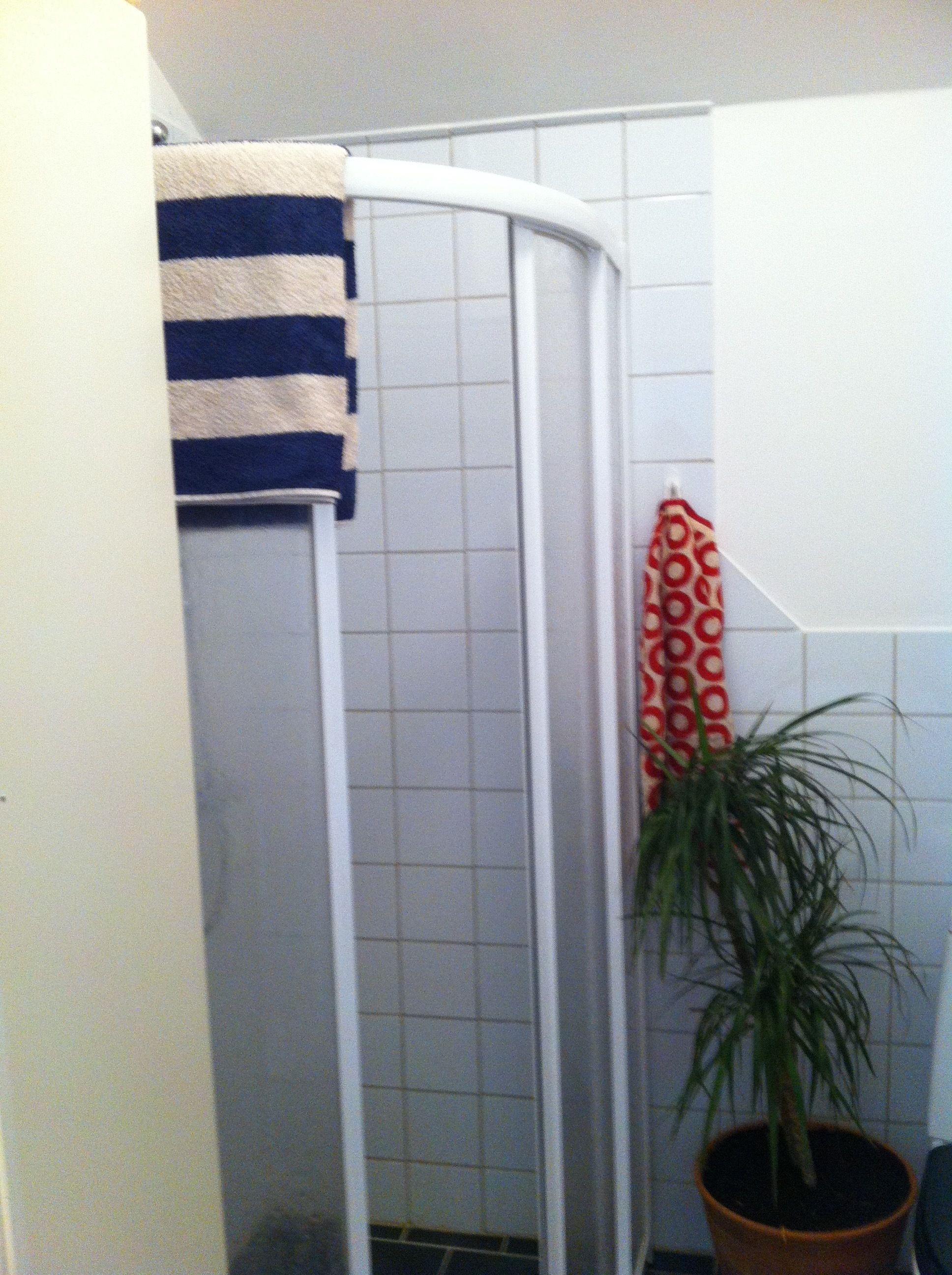 Ein Duschbereich mit einer weiß gefliesten Wand, einem blau-weiß gestreiften Handtuch und einem rot-weißen gemusterten Handtuch, das an der Wand hängt.