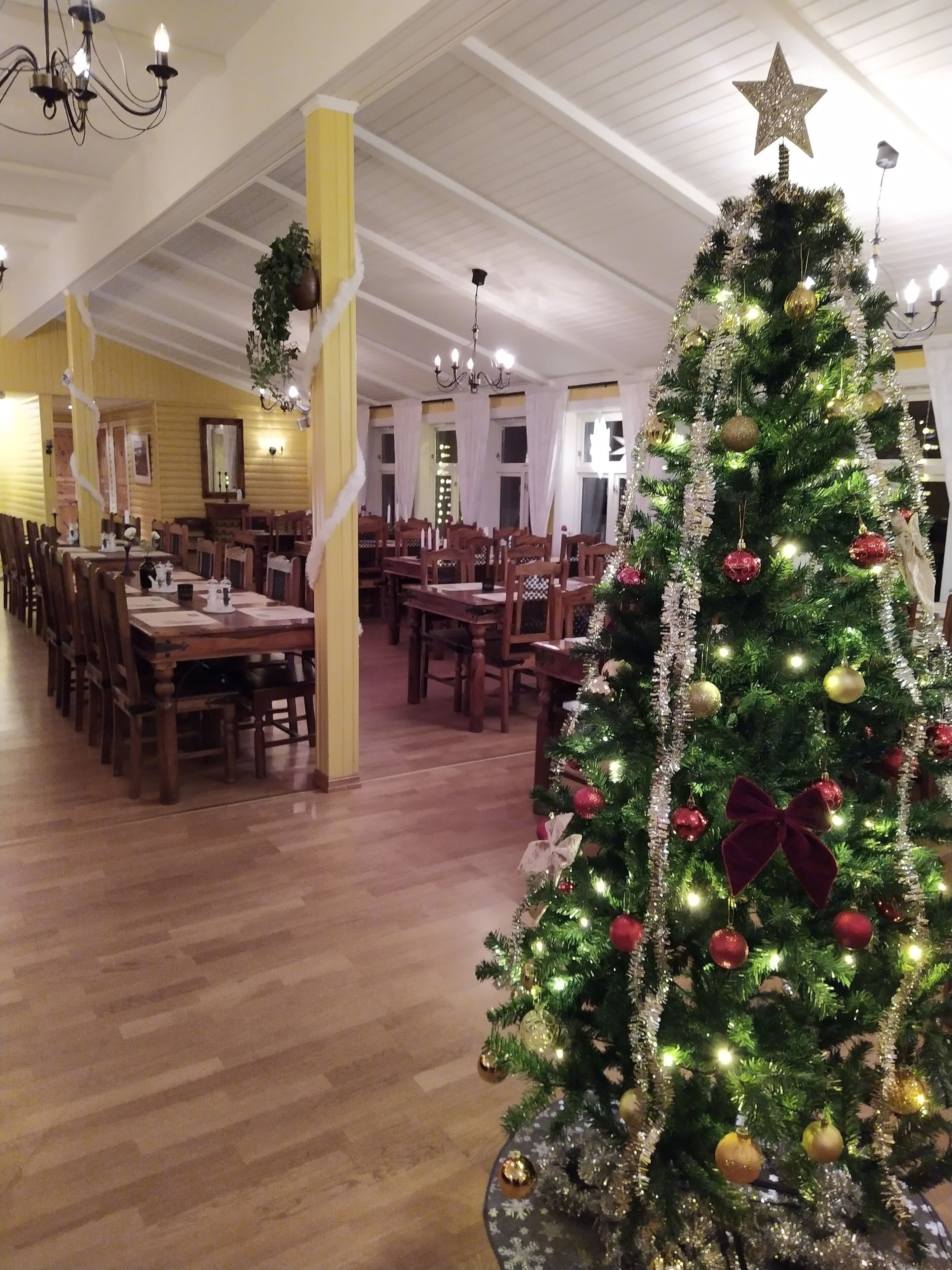 Uma sala de jantar com mesas e cadeiras de madeira, uma árvore de Natal decorada e um lustre.