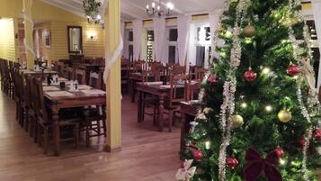 Uma sala de jantar com mesas e cadeiras de madeira, uma árvore de Natal decorada e um lustre.