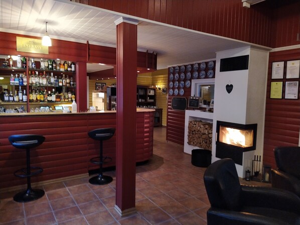A bar area with a wooden bar counter, multiple stools, and a variety of liquor bottles. There is a fireplace with a log fire, and a wall decorated with framed certificates and a heart-shaped plaque. - Lyngseidet Gjestegård (Lyngen)