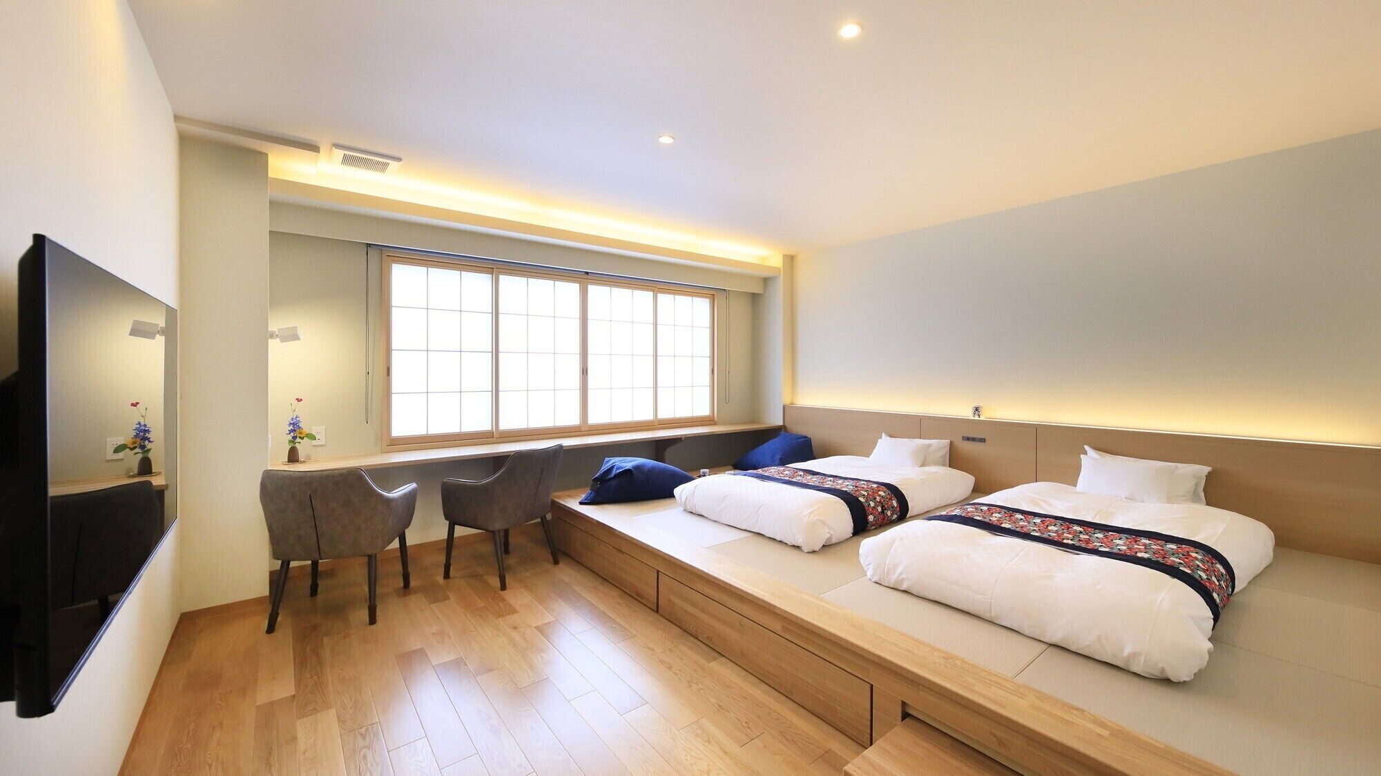 A hotel room with a bed, bedside tables, a desk, chairs, a large window, and a TV.