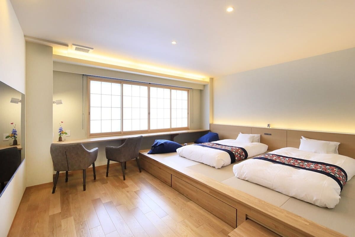 A hotel room with a bed, bedside tables, a desk, chairs, a large window, and a TV.