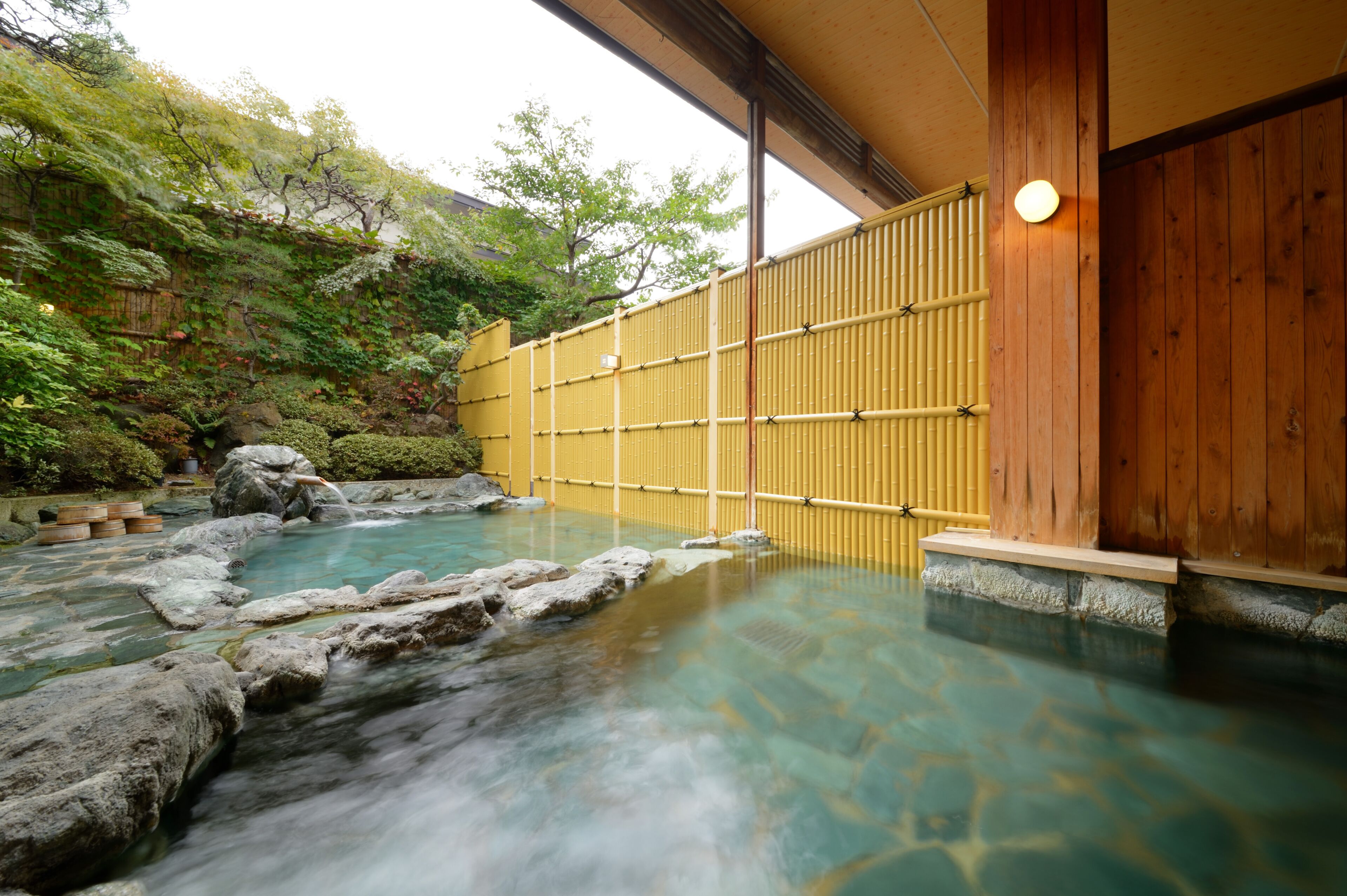 A traditional Japanese onsen with a wooden bathhouse, a natural rock pool, and a bamboo fence.