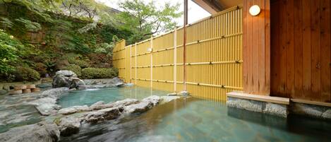 A traditional Japanese onsen with a wooden bathhouse, a natural rock pool, and a bamboo fence.