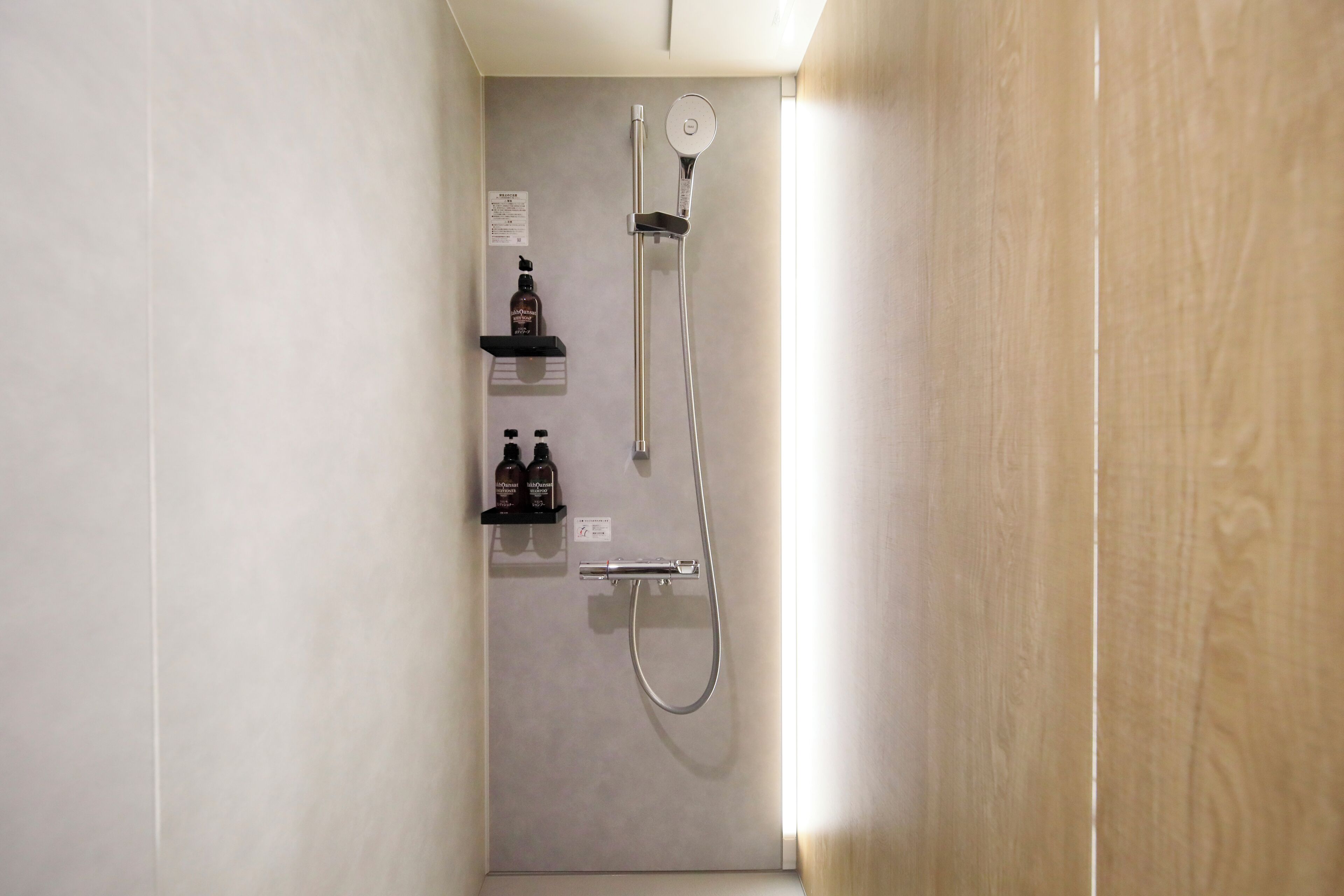 Un espacio de ducha estrecho con un cabezal de ducha de mano y un grifo instalado en la pared. Hay dos botellas en un estante sobre la ducha.