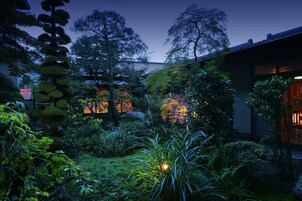 Un jardín tranquilo con diversos árboles y plantas, un césped bien cuidado y un edificio con ventanas iluminadas al fondo.