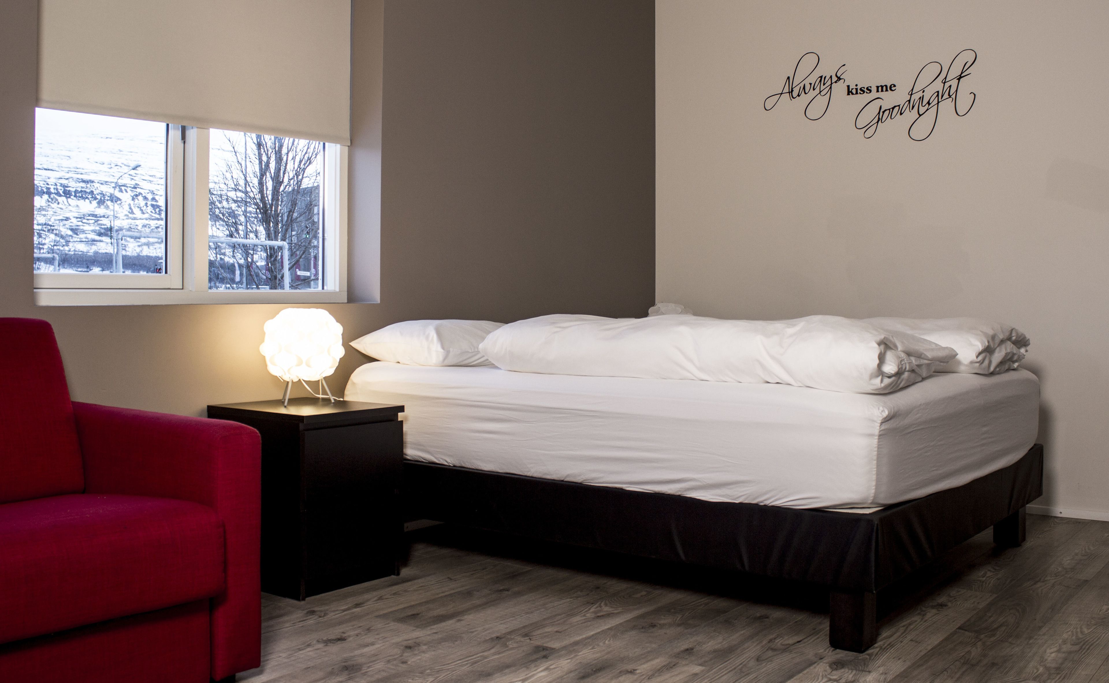 Un dormitorio con cama, una mesita de noche con lámpara, una silla roja y una ventana con vista a árboles.