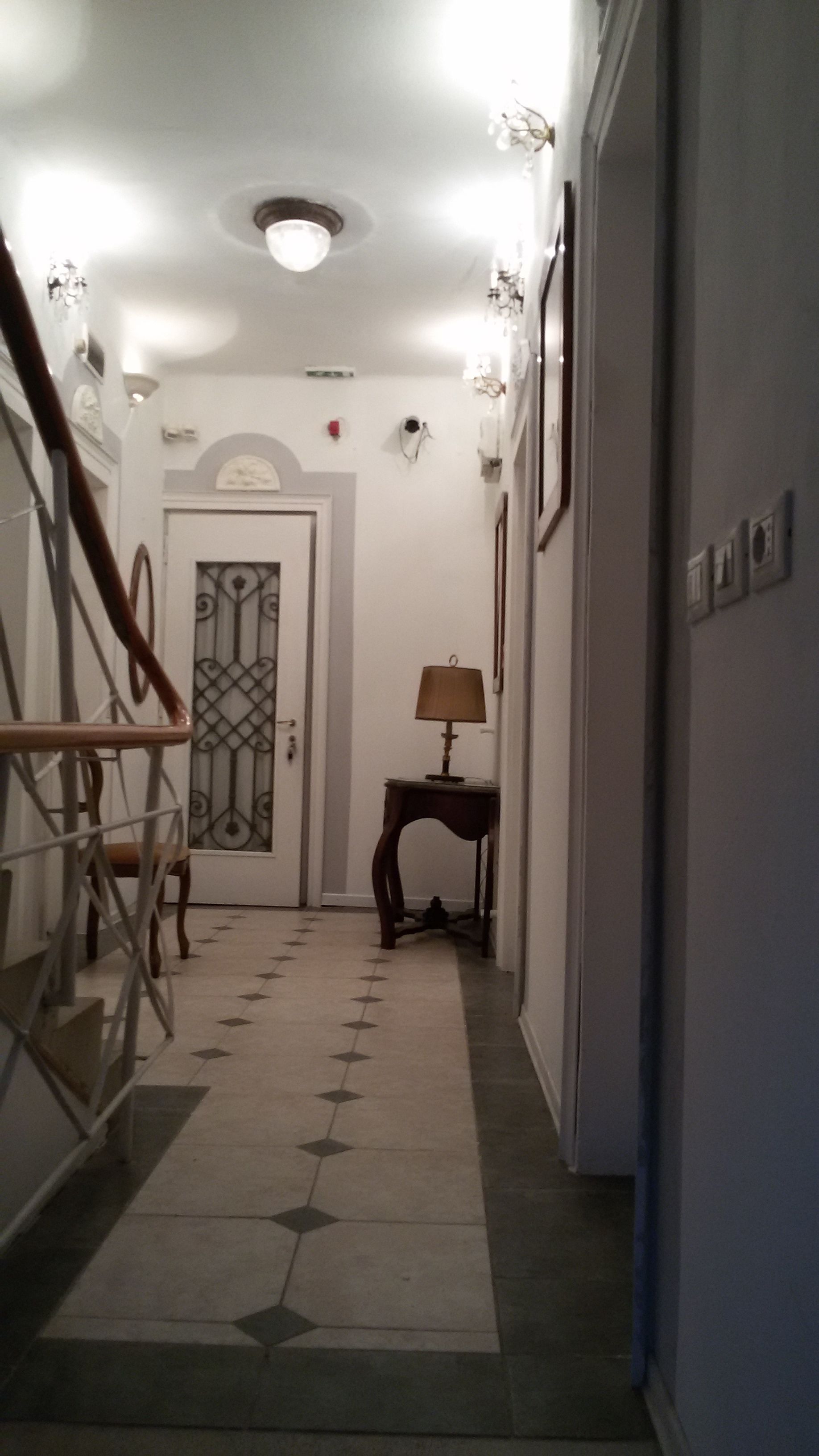 A hallway with a patterned floor, a door with a decorative glass panel, a small table with a lamp, and a staircase with wooden railings.
