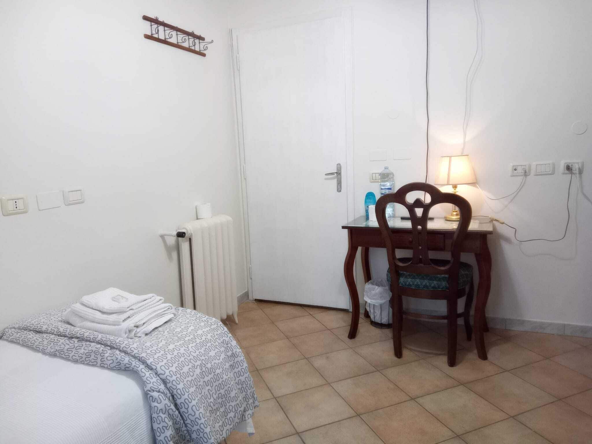 A small, clean room with a bed, a desk, and a chair. There is a radiator, a lamp, and a few items on the desk.