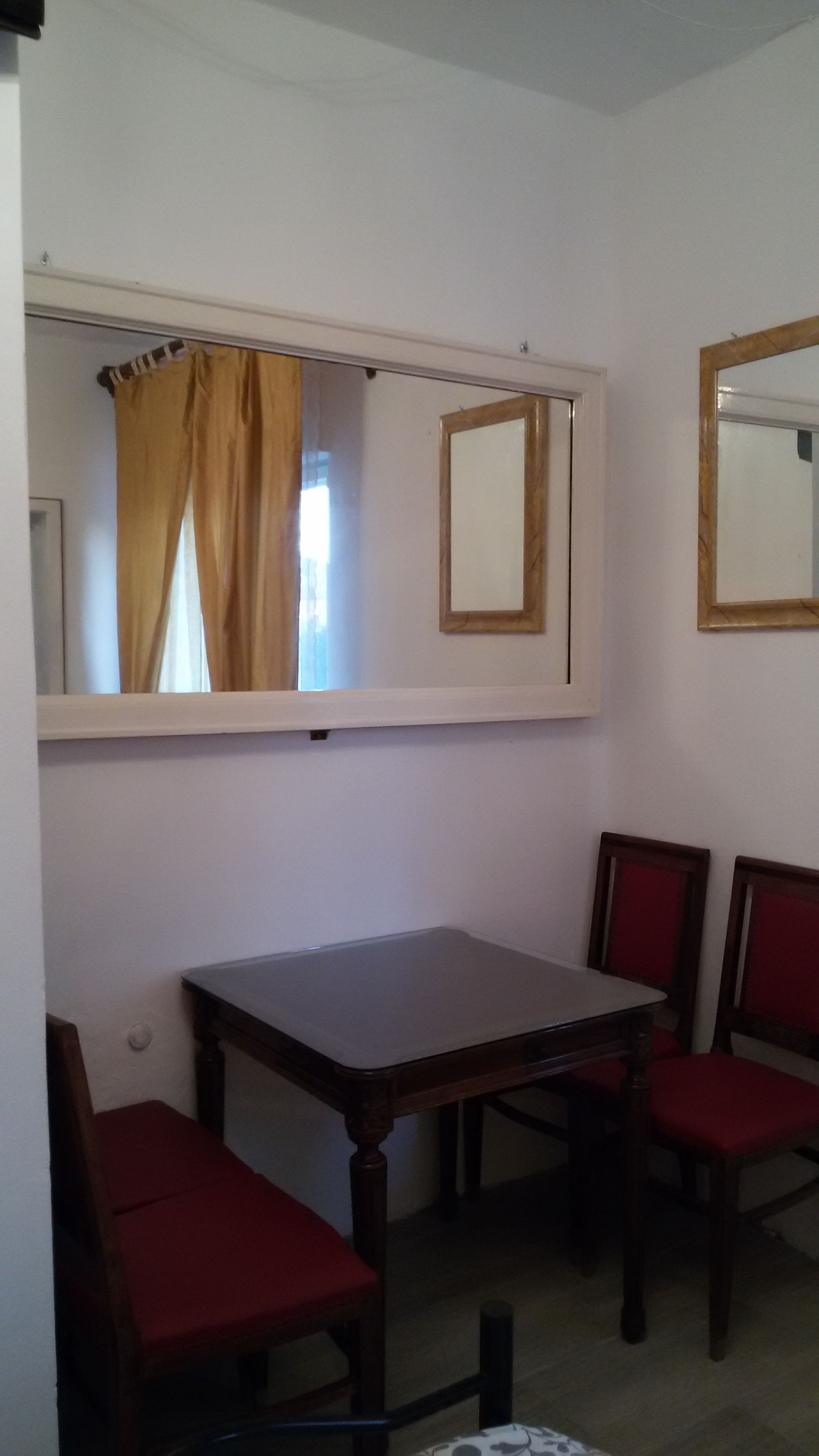 Une petite pièce avec une table en bois et deux chaises rouges, un miroir au mur et une fenêtre avec un rideau.