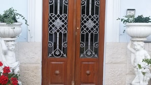 A wooden double door with decorative ironwork, flanked by two white statues, and a welcome mat at the entrance. - Villa Fiorita (Monfalcone)