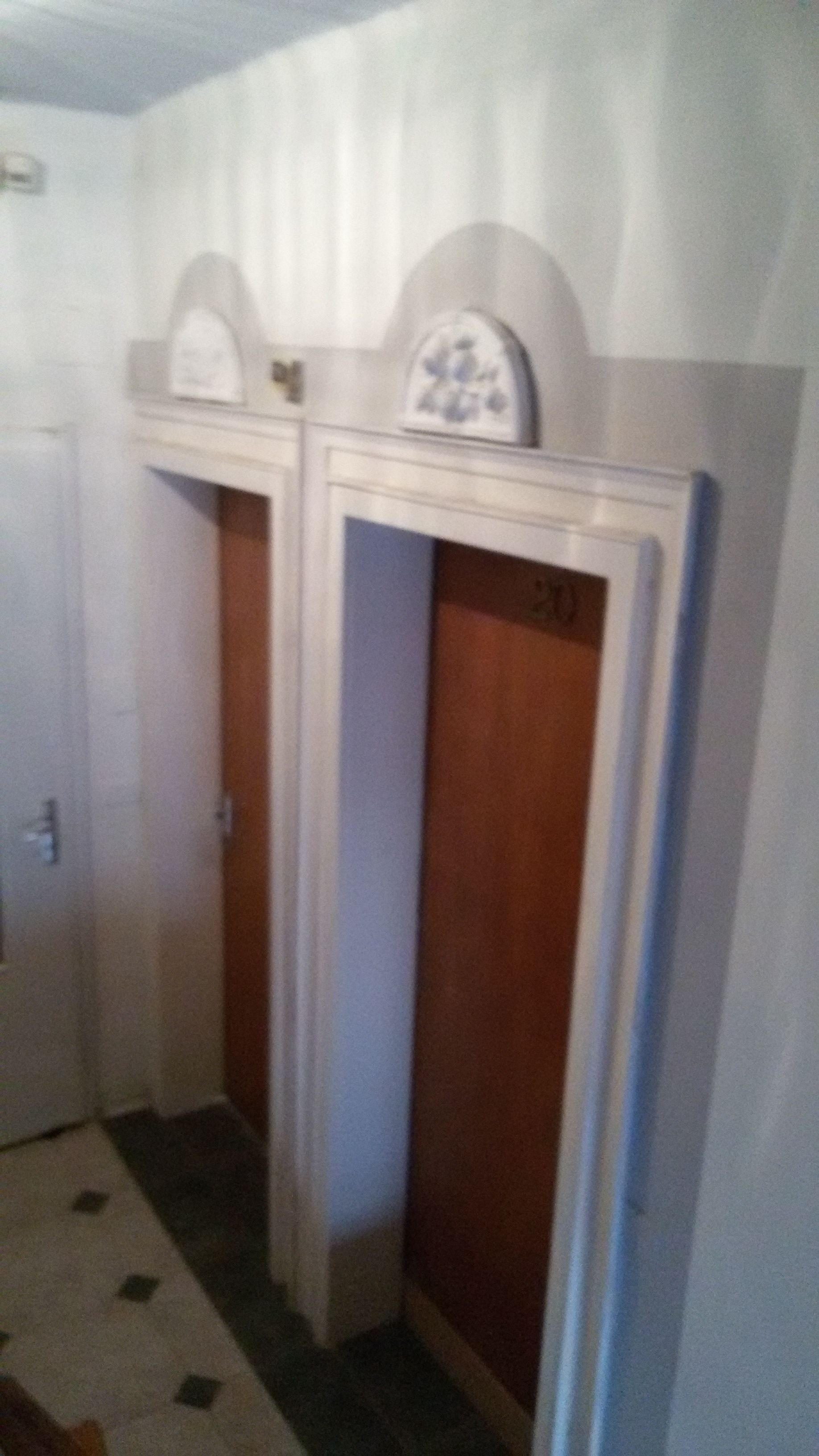 A hallway with two wooden doors, one of which has a number '20' on it. There is a white wall with a decorative element above the doors.