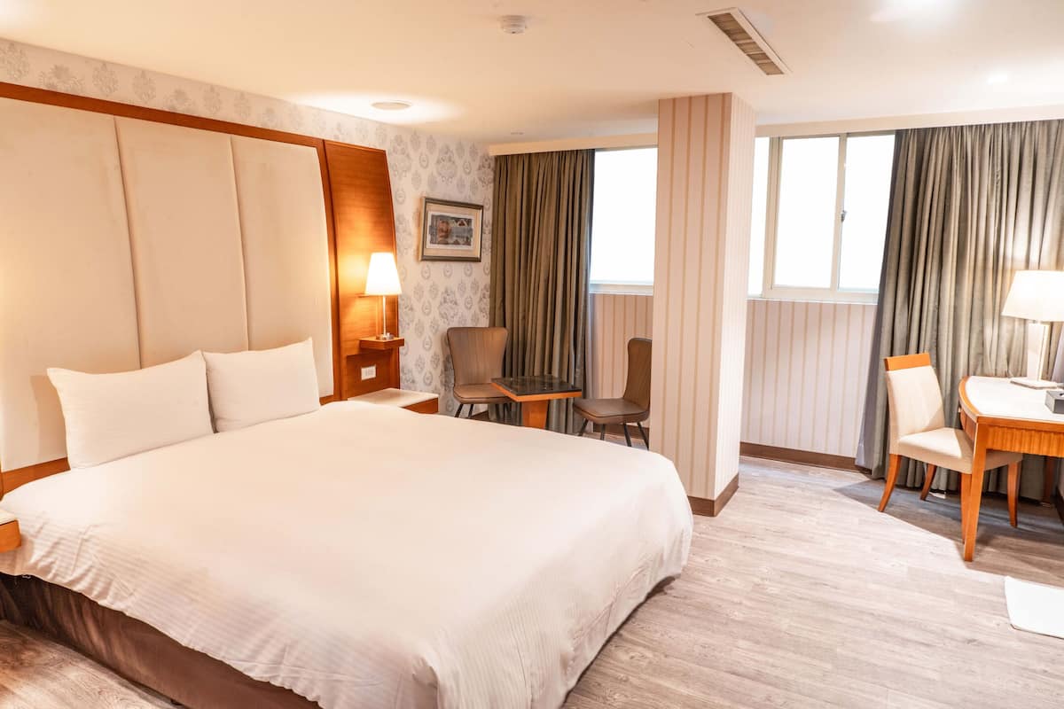 A hotel room with a large bed, a desk, two chairs, a window with curtains, and a wall-mounted picture.