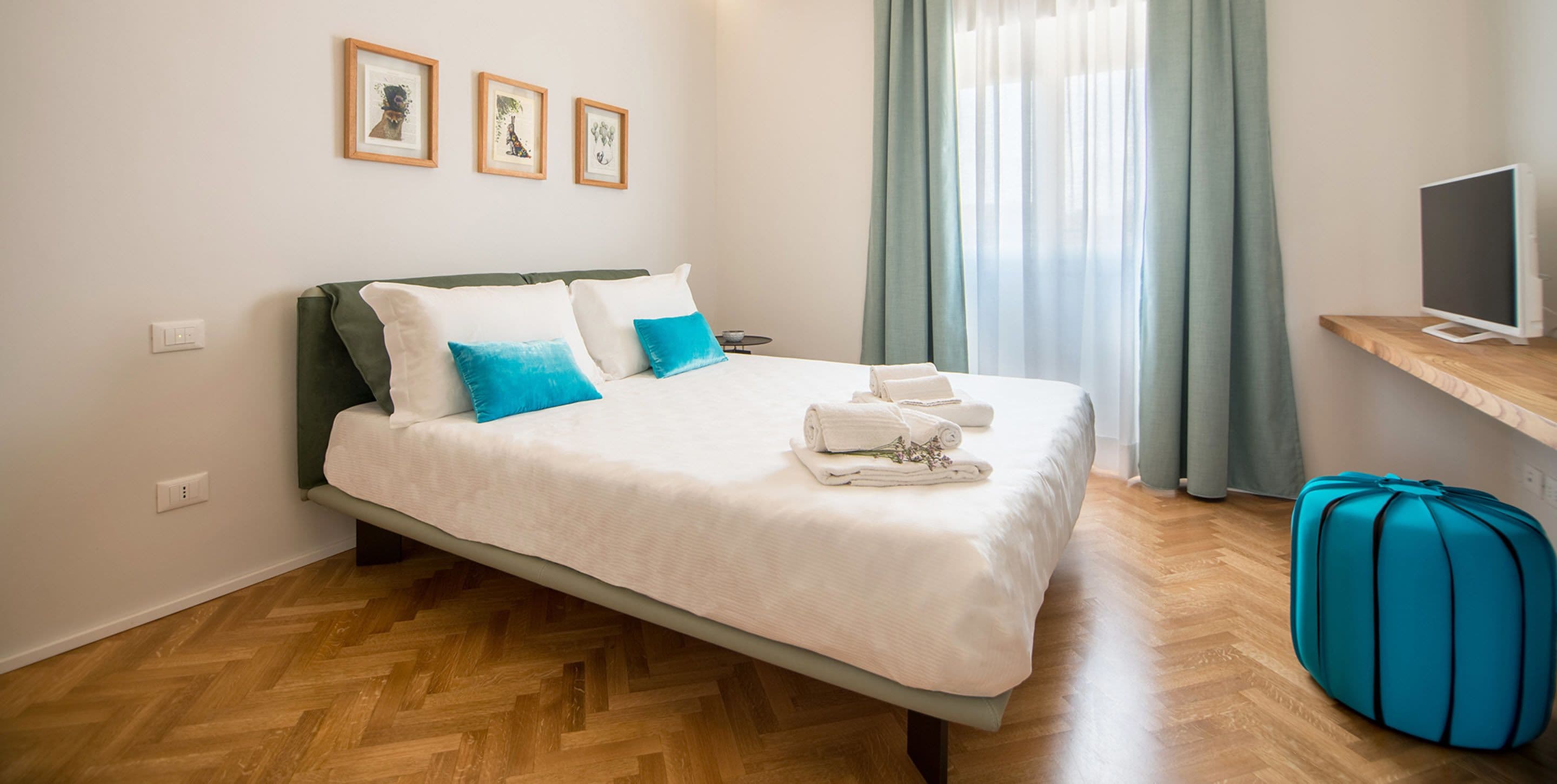 Ein ordentlich bezogenes Bett mit weißer und blauer Bettwäsche, ein Holzboden, ein blauer Koffer und ein Fernseher auf einem Holzschrank.
