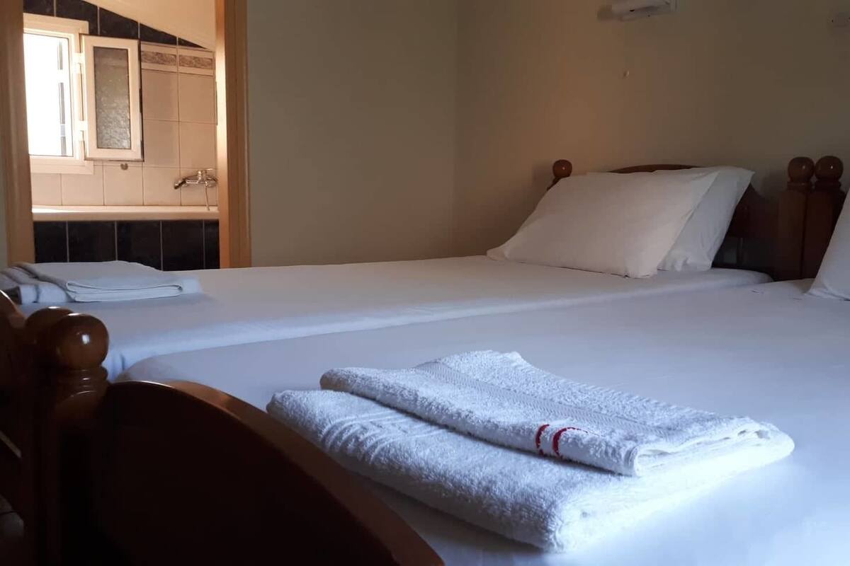 A hotel room with a bed, two pillows, a wooden headboard, a wooden chair, and a bathroom visible through an open door.