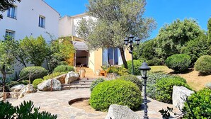 A hotel with a stone pathway leading to the entrance, surrounded by well-maintained greenery and a clear blue sky. - Hôtel Cala di l'Oru (L'Île-Rousse)