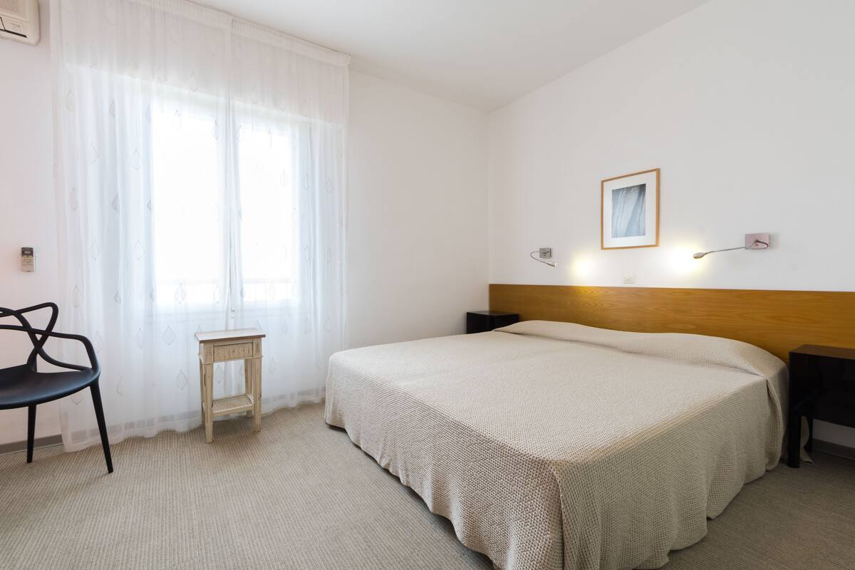 Habitación de hotel con una cama grande, una mesita pequeña, una silla y una lámpara fijada en la pared.