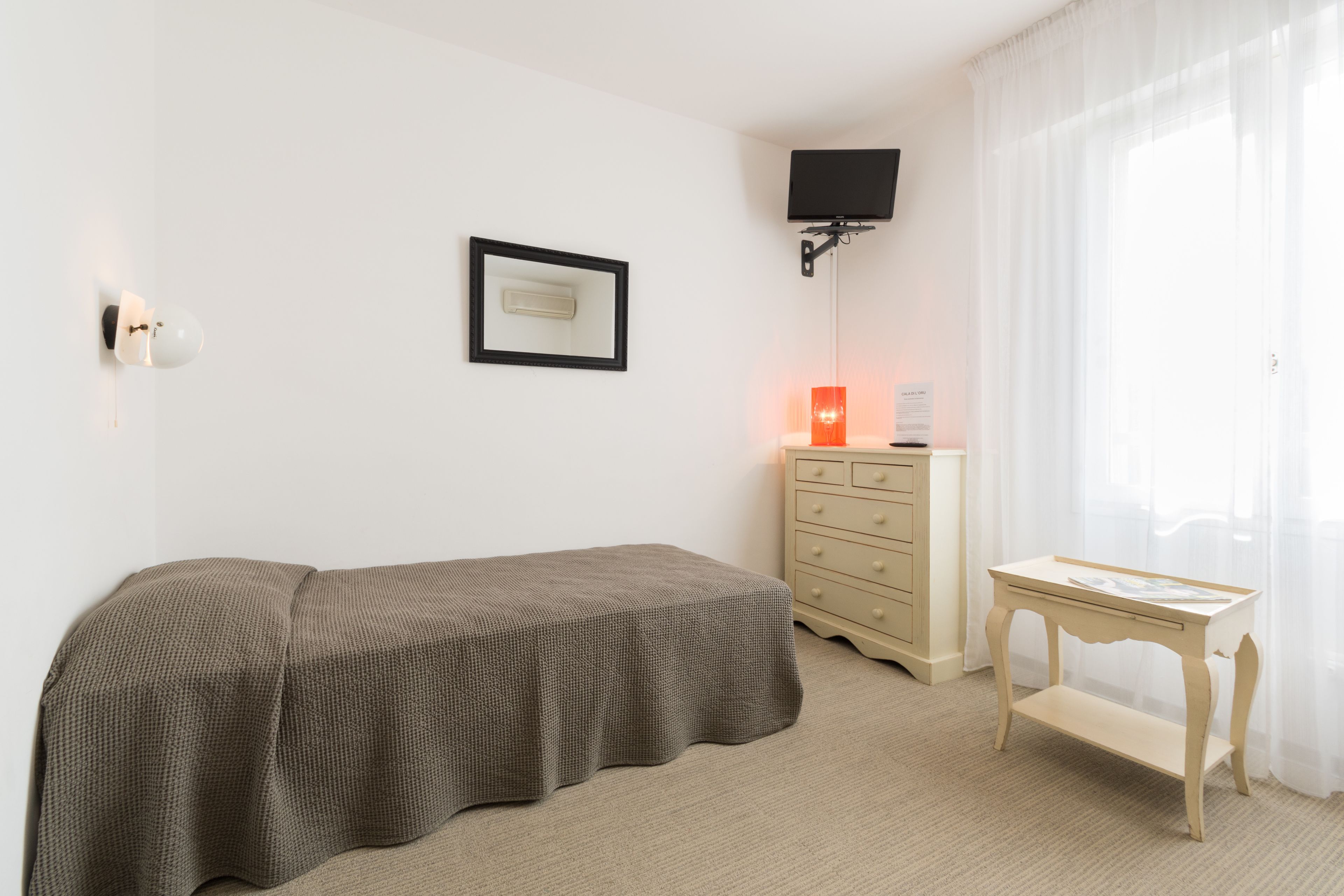 Ein Schlafzimmer mit Bett, Kommode, Nachttisch, einem an der Wand montierten Fernseher und einem Fenster mit durchsichtigen Vorhängen.