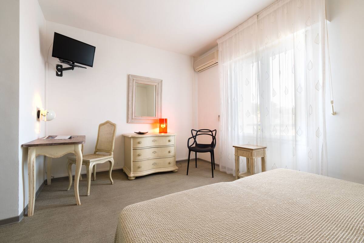 Una habitación de hotel con cama, escritorio, silla, cómoda, televisor montado en la pared y ventana con cortinas translúcidas.