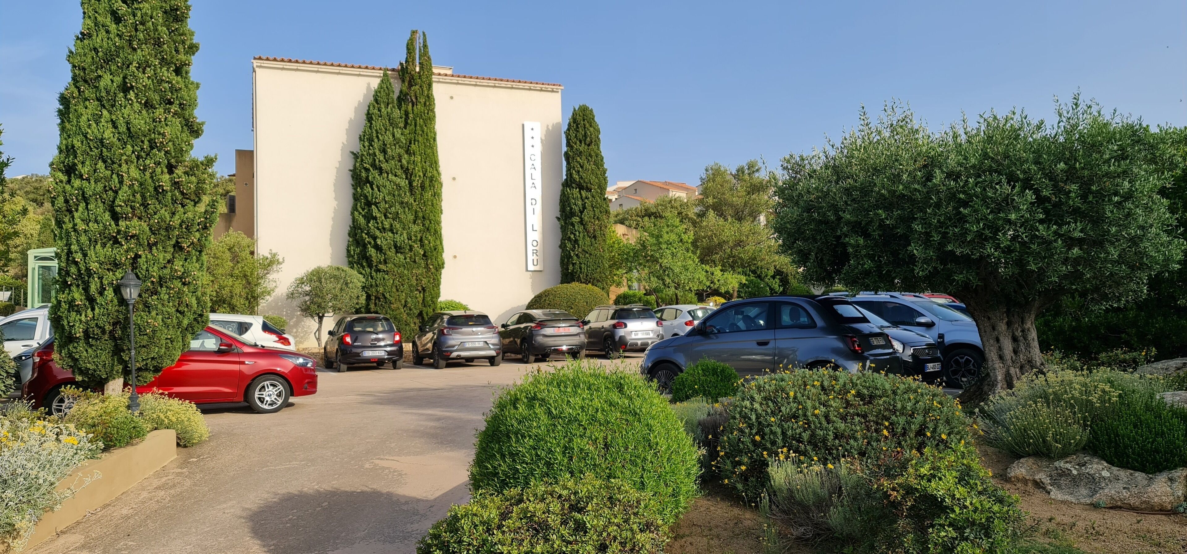 Zona d'aparcament amb diversos cotxes, un edifici amb un rètol i vegetació als voltants.