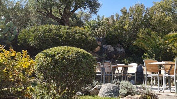 An outdoor seating area with tables and chairs, surrounded by various plants and trees. - Hôtel Cala di l'Oru (L'Île-Rousse)