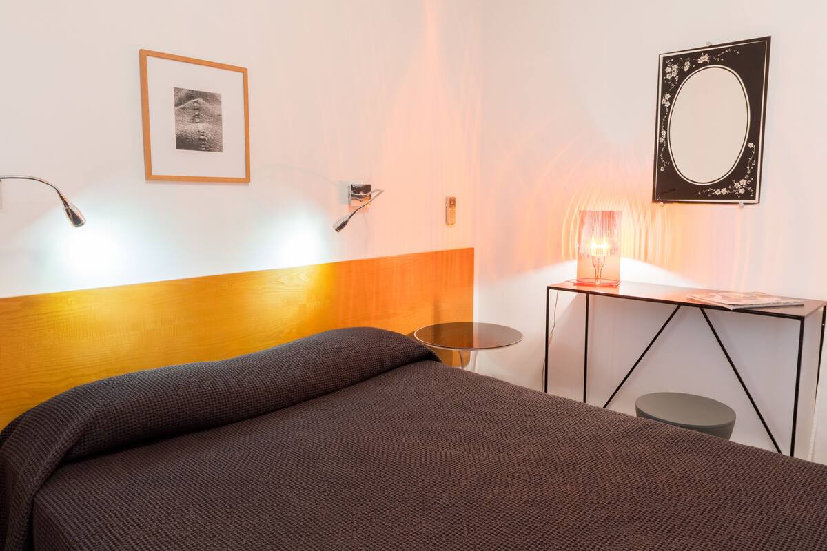 Habitación de hotel moderna con cabecera de madera, una cama individual, una mesita redonda pequeña, un escritorio con espejo y un cuadro en la pared.