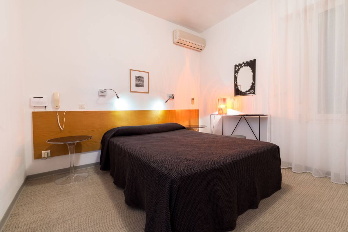 Habitación de hotel con cama, mesita de noche, lámpara y unidad de aire acondicionado.