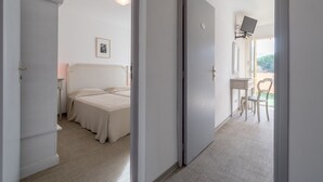 A bedroom with a bed, a bedside table, a chair, and a desk with a chair. There is a door leading to another room. - Hôtel Cala di l'Oru (L'Île-Rousse)