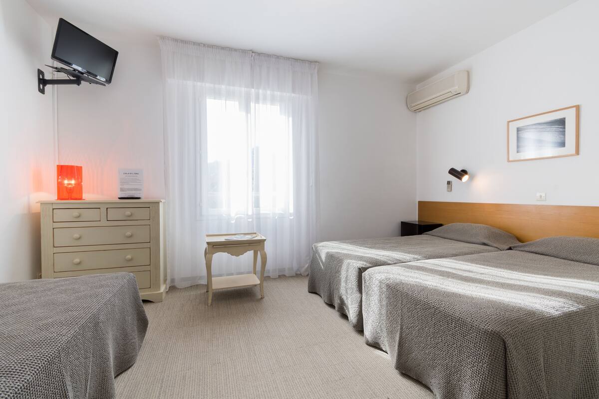 Habitación de hotel con dos camas, un televisor montado en la pared, una mesita con lámpara y una ventana con cortinas translúcidas.