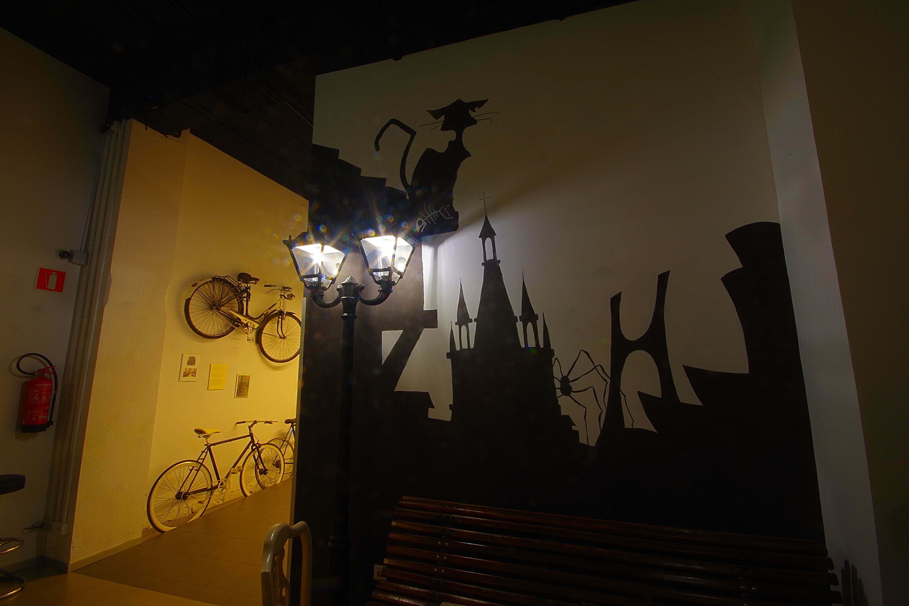 Un couloir avec des vélos, un banc et une fresque murale représentant un chat et le panorama d’une ville.