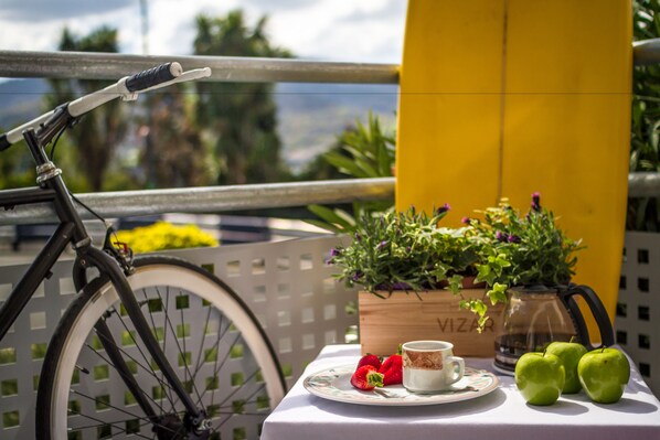 A bicycle, a table with a plate of strawberries, a cup, and green apples on a balcony with a yellow column and a potted plant. - All Iron Hostel (Bilbao)