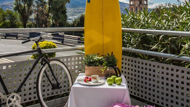 A yellow surfboard, a bicycle, a table with food, and a basket on a balcony with a view of a church and mountains. - All Iron Hostel (Bilbao)