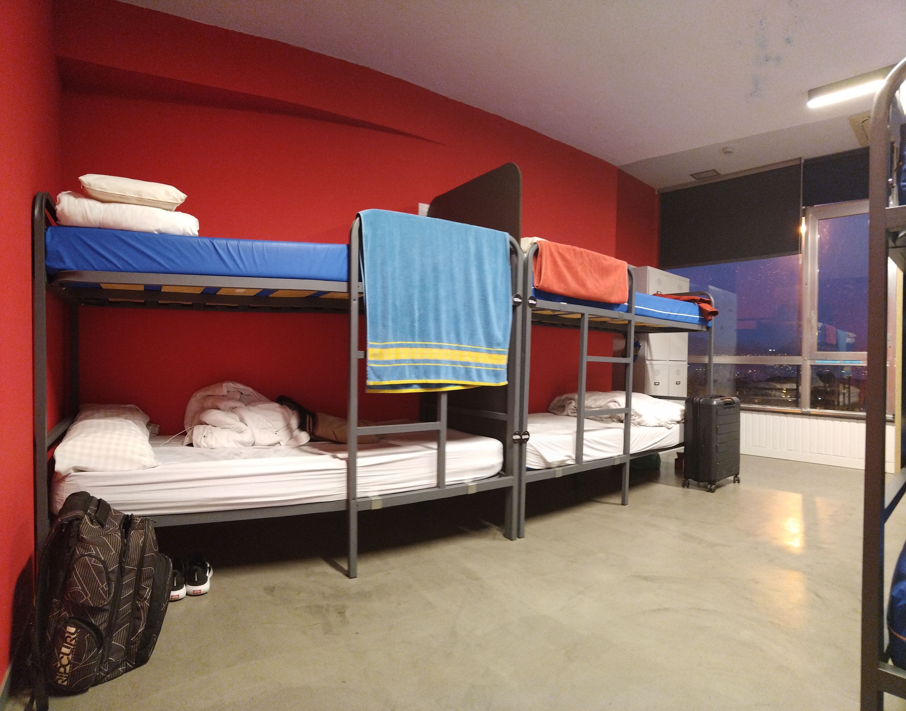 A bunk bed room with a red wall, a window with a view, and a suitcase on the floor.