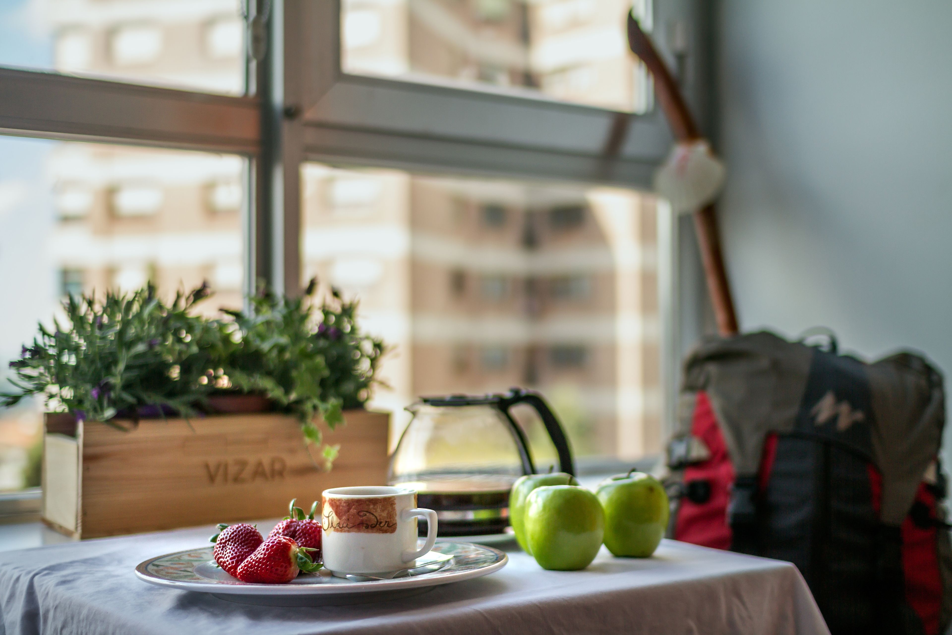 A table with a cup, strawberries, and green apples, a wooden box labeled 'VIZAR', and a window with a view of buildings.
