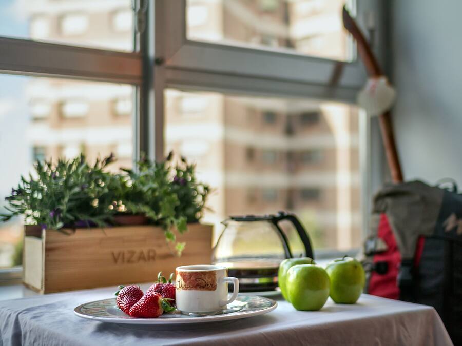 Una mesa con una taza, fresas, manzanas verdes, una caja de madera con la etiqueta 'VIZAR', y una ventana con vistas a edificios.