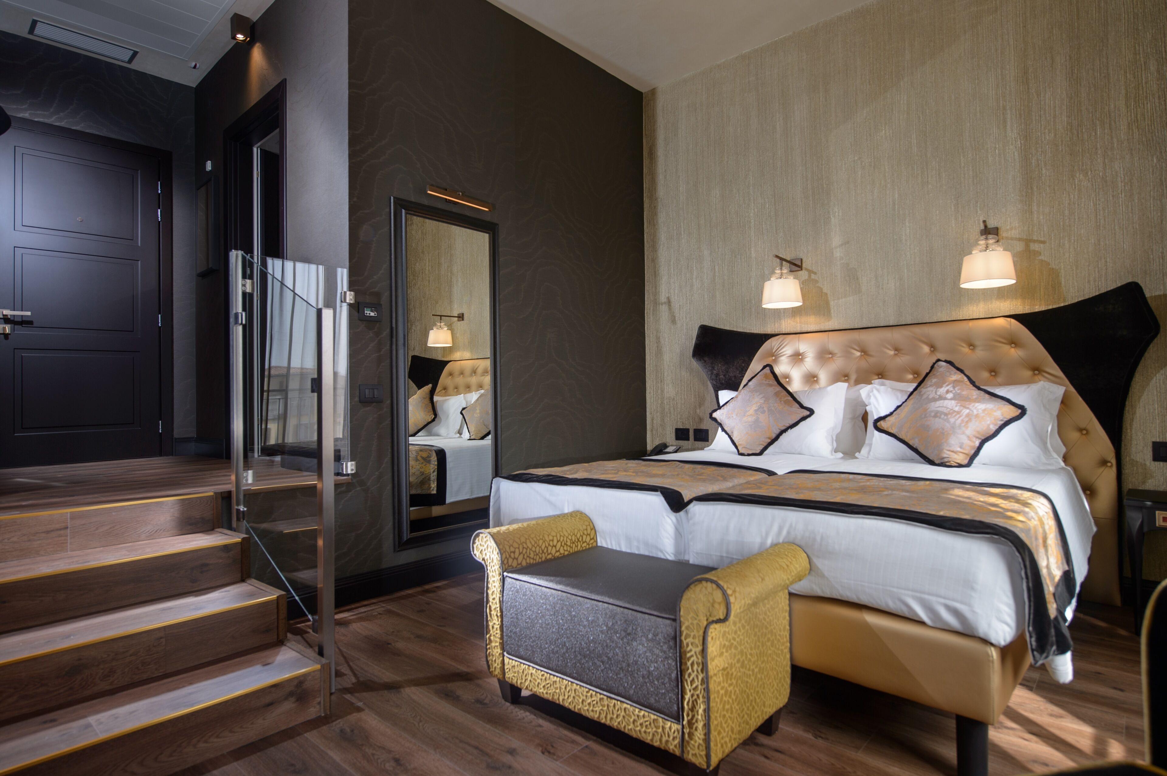 A modern hotel room with a large bed, a sitting area, and a staircase leading to an entrance.