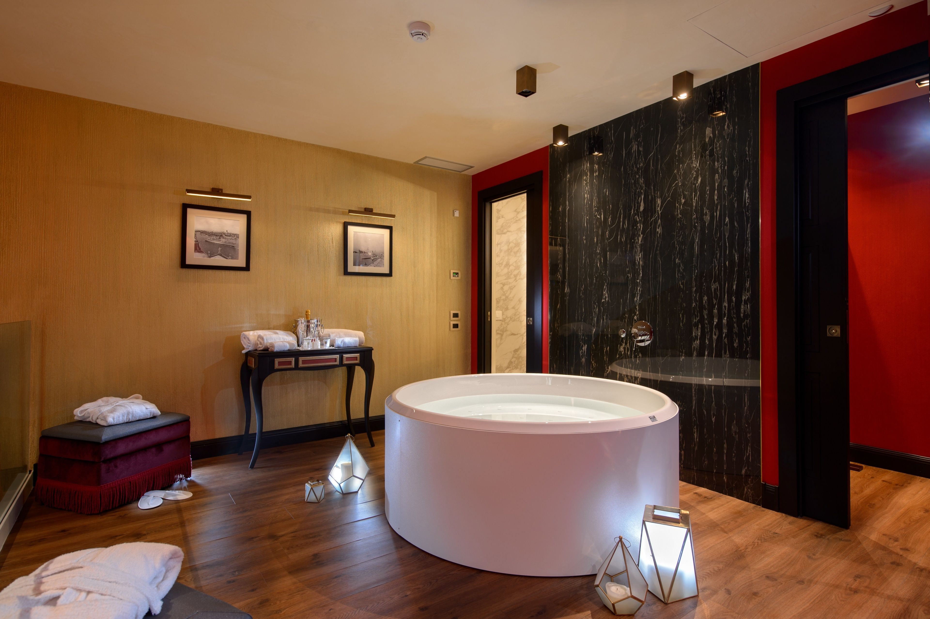 A modern spa room with a large white bathtub, wooden flooring, and a red accent wall.
