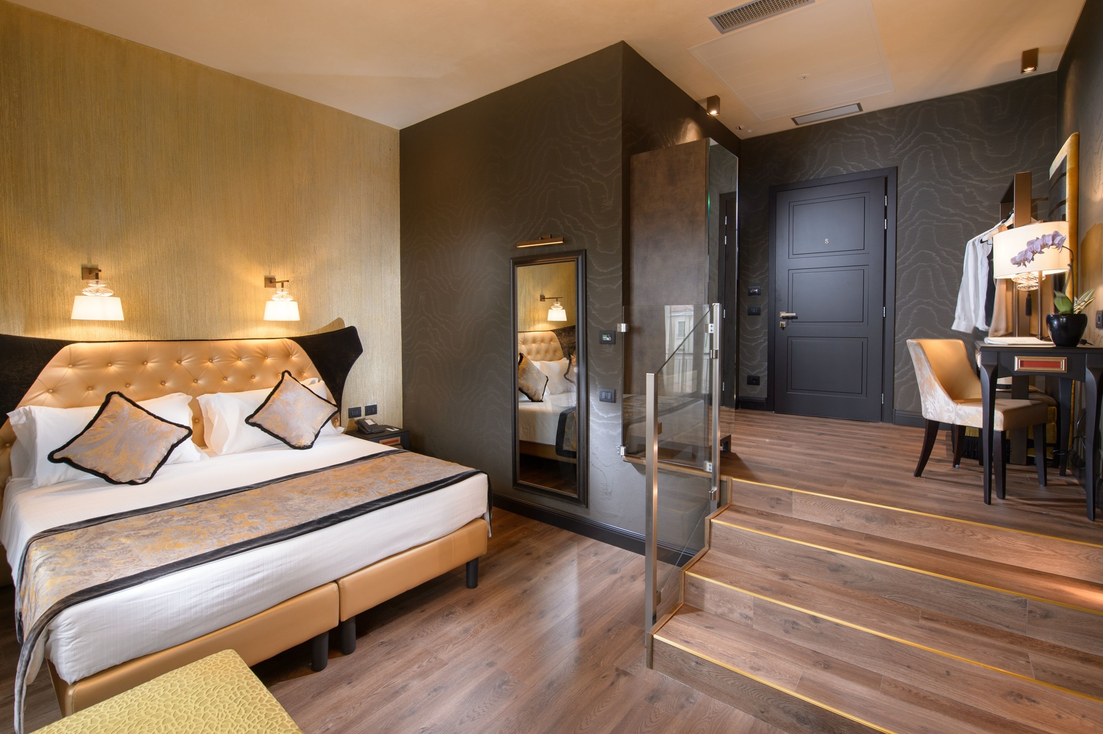 A modern hotel room with a large bed, a sitting area with a chair, a desk, and a mirror.