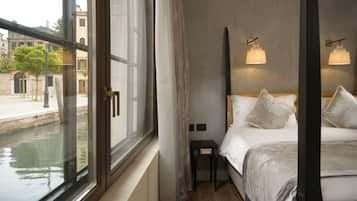 Hotellihuone, jossa on suuri ikkuna, josta avautuu näkymä kanavalle, valkoisilla lakanoilla varustettu sänky, pieni yöpöytä ja kaksi seinälle asennettua lamppua.