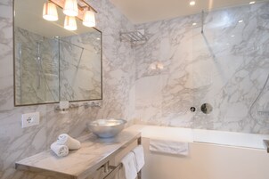 Un baño moderno con pared de mármol, un espejo grande, ducha con cerramientos de vidrio y una bañera blanca.