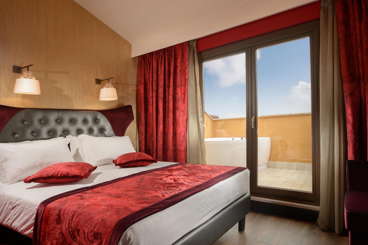 Ein Hotelzimmer mit einem großen Bett, roter Bettwäsche, zwei Wandlampen und Blick auf den Balkon.