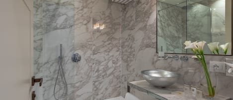Un baño moderno con pared de ducha de mármol, bañera con cerramiento de vidrio, lavabo tipo cuenco grande y espejo con iluminación superior.