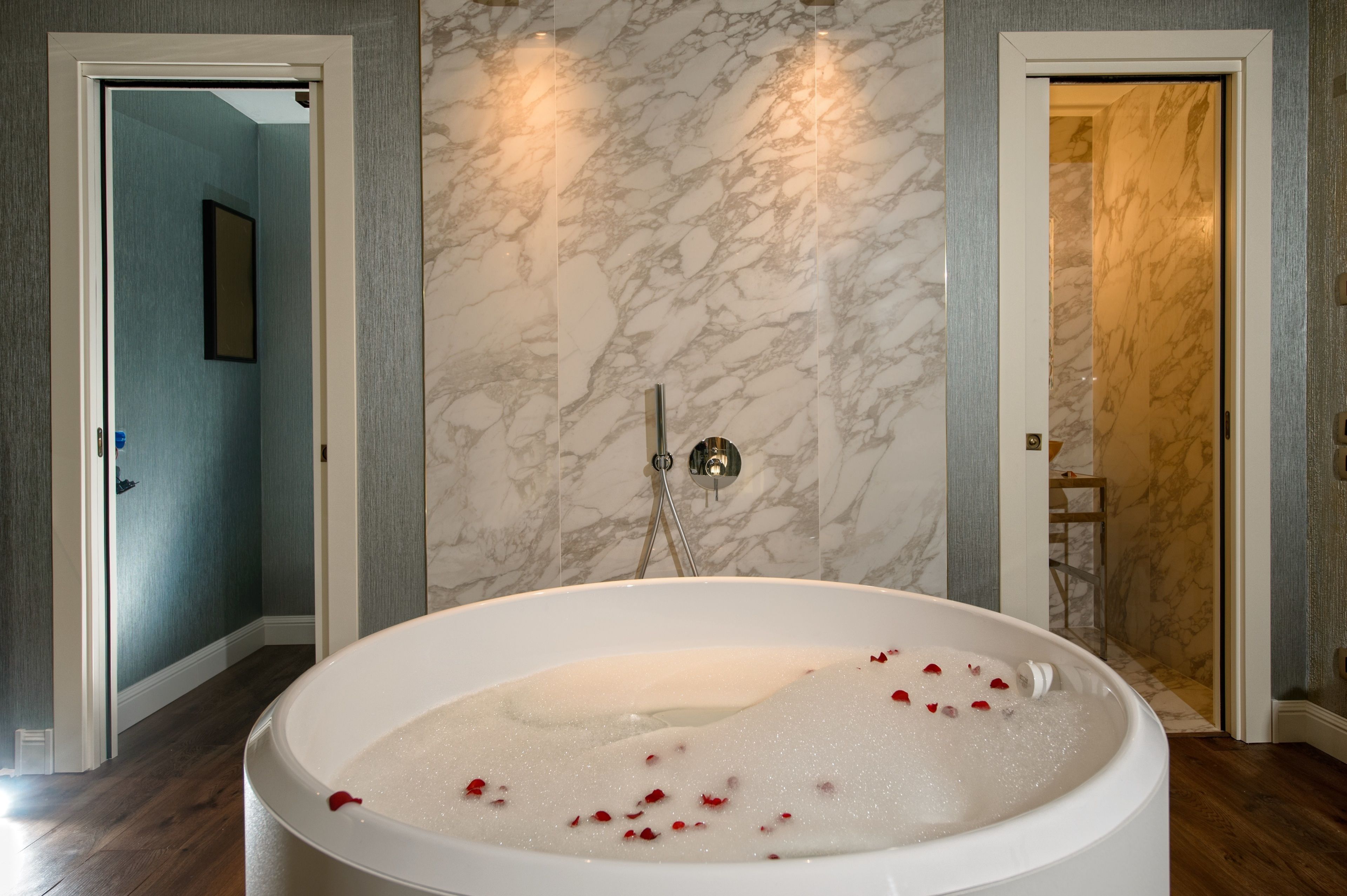 A spacious bathroom with a large bathtub filled with water and rose petals, marble walls, and a shower area.