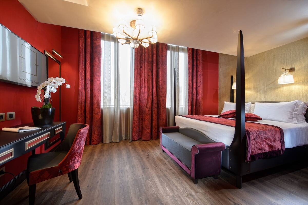 Ein Hotelzimmer mit Himmelbett, Schreibtisch mit Stuhl, Fernseher und großem Fenster mit roten Vorhängen.