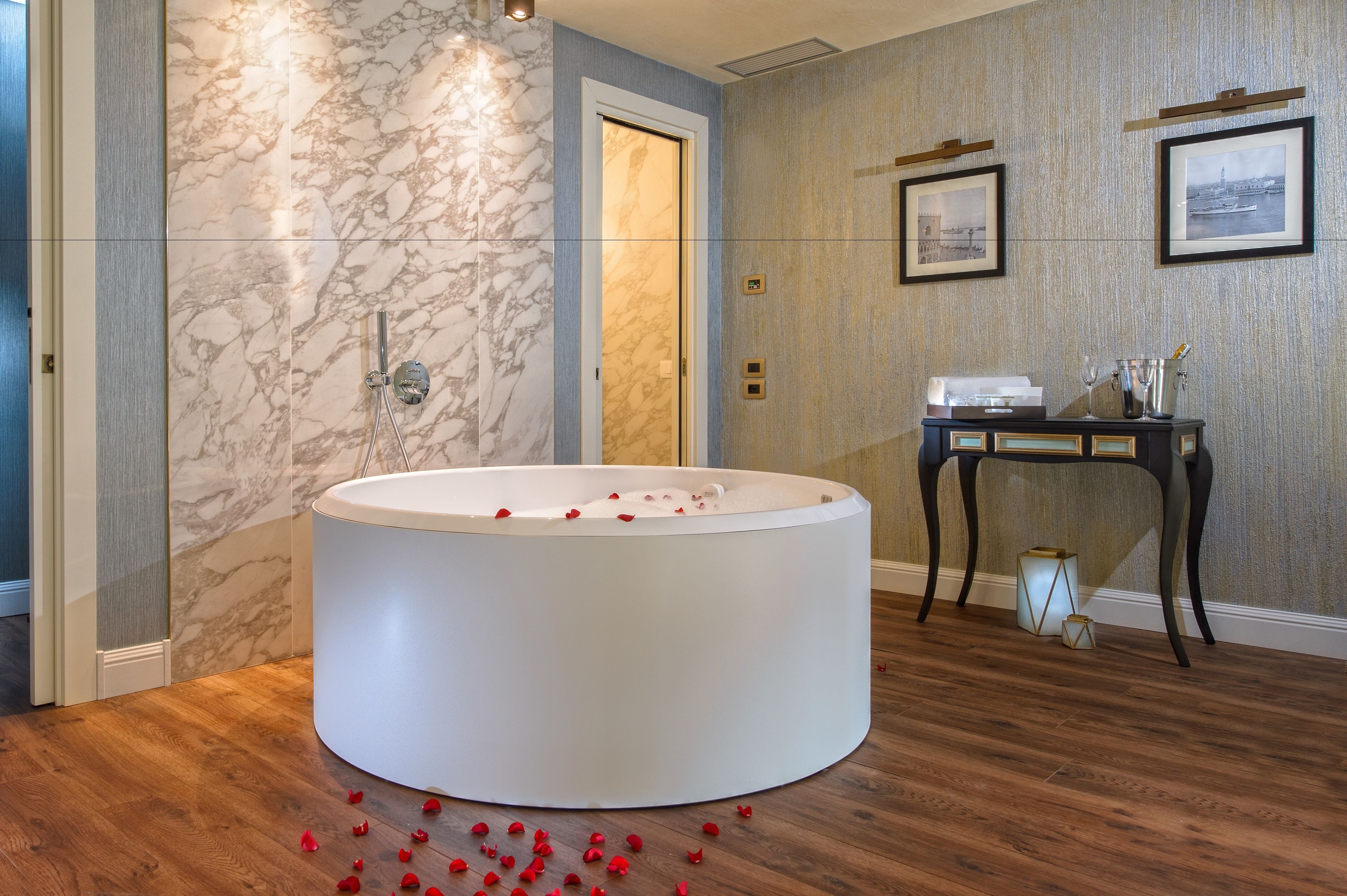 A modern bathroom with a large bathtub, marble walls, wooden flooring, and a small table with toiletries.