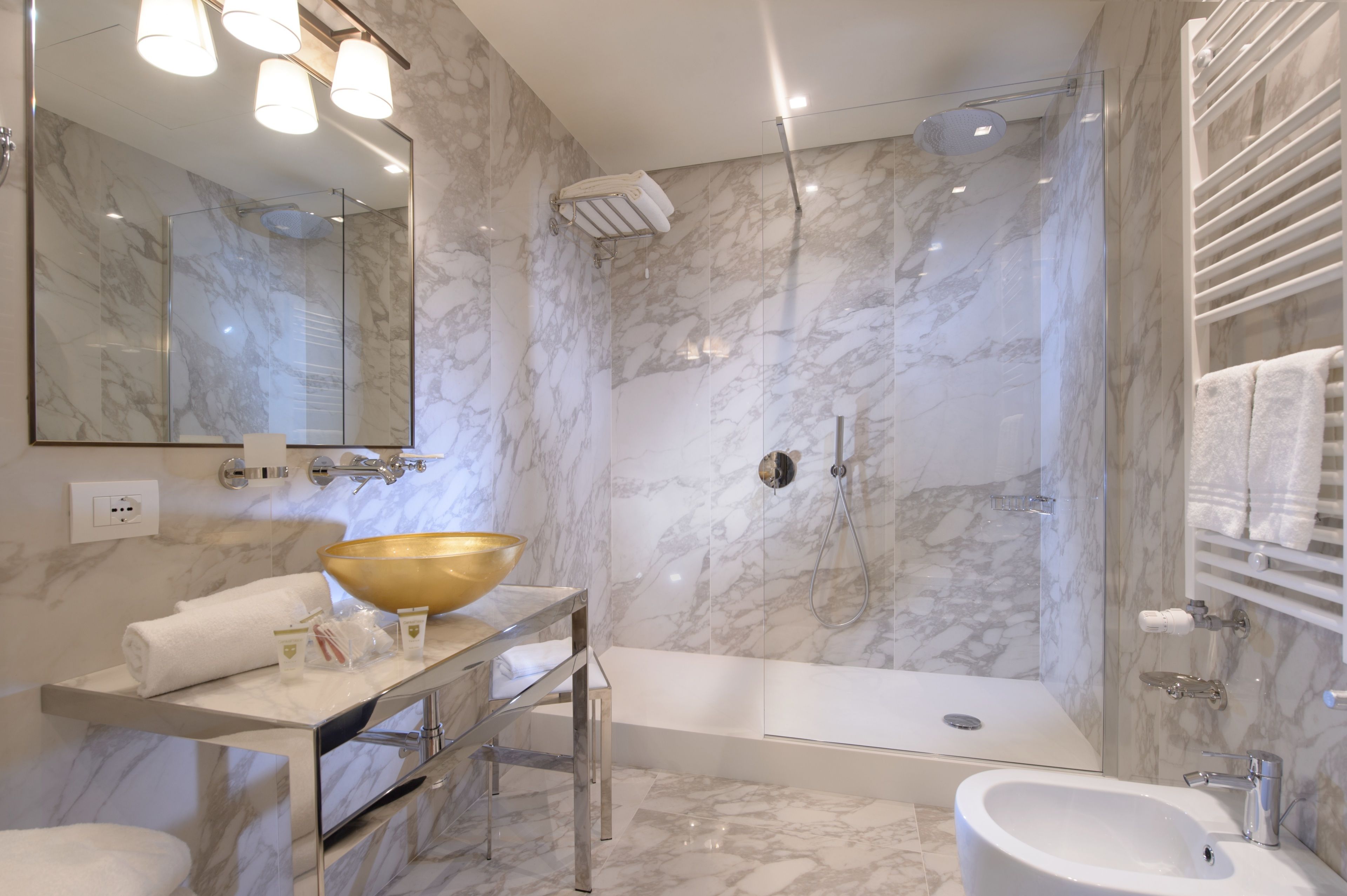A modern bathroom with a marble wall, a large mirror, a gold basin, a bathtub, and a shower area.