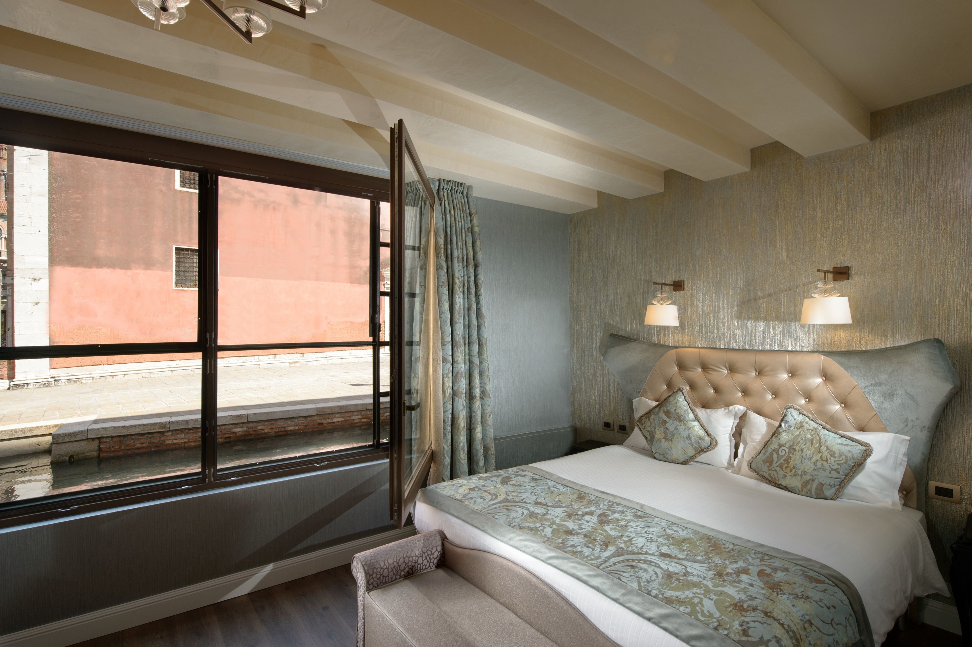 Un dormitorio con una cama grande, una ventana con cortinas, un cabecero con luces fijadas a la pared y vista a la calle.